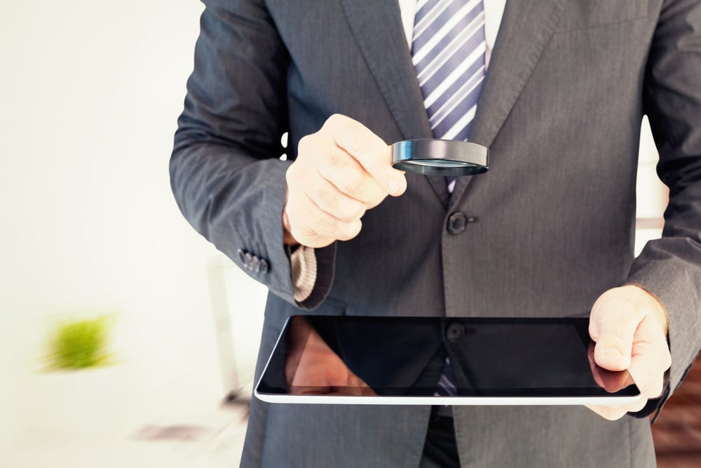 Businessman looking at tablet with magnifying glass against laptop on desk with glasses and notepad