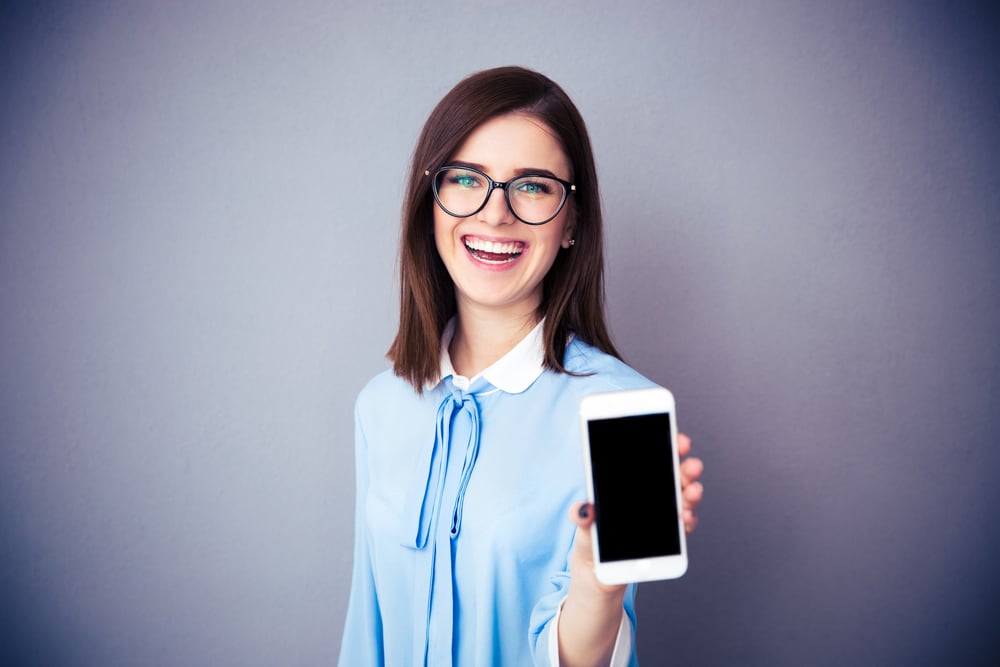 Laughing businesswoman showing blank smartphone screen over gray background. Wearing in blue shirt and glasses. Looking at camera.