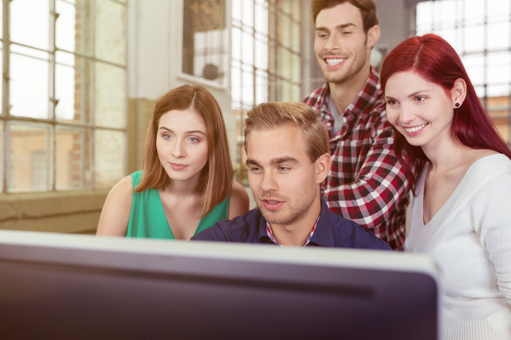 Informal young business team anxiously watching the success of their project as they stand grouped around a large screen desktop computer